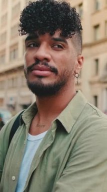 VERTICAL VIDEO: Young happy man with beard dressed in an olive-colored shirt stands next to the road, turns his head, crosses his arms and looks at the camera