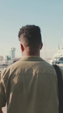 VERTICAL VIDEO: Young man dressed in an olive-colored shirt is standing in the port looking at the yachts. Back view