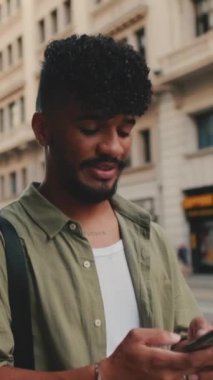 VERTICAL VIDEO: Young happy man with beard dressed in an olive-colored shirt walks through the center of the old city using mobile phone