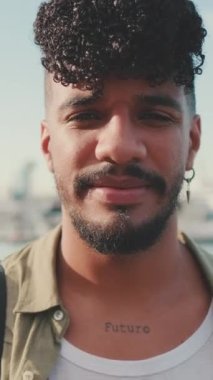 VERTICAL VIDEO: Close-up of young man dressed in an olive color shirt sitting on the waterfront in the port looking at the camera