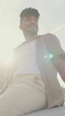 Zeytin rengi tişört giymiş genç bir adam limandaki gezinti güvertesinde oturur ve yatlara bakar.