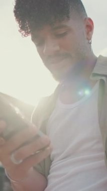 VERTICAL VIDEO: Close up, young man dressed in an olive-colored shirt is sitting on the waterfront in the port watching videos and photos on his cellphone. Backlight