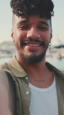 VERTICAL VIDEO: Close up, young smiling man dressed in an olive color shirt makes video call while standing on the waterfront in the port