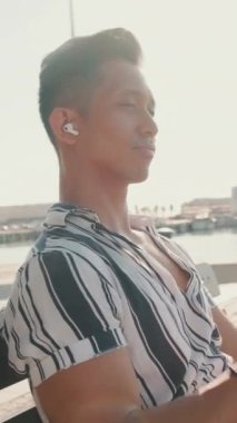 VERTICAL VIDEO: Young man tourist sits on bench on the pier, listens to music. Backlight