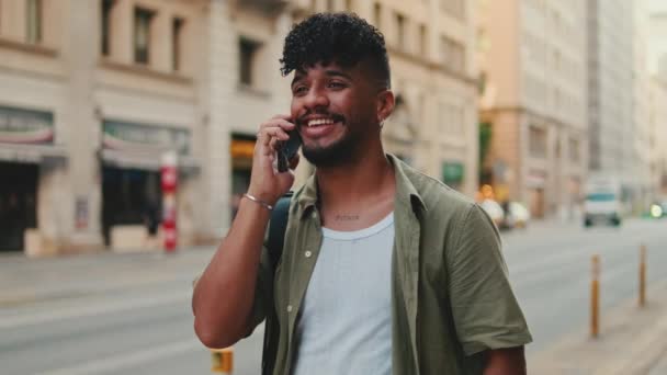 オリーブ色のシャツに身を包んだ若い幸せな男は 古い街の背景の道路の横に立っている間 携帯電話で話しています — ストック動画
