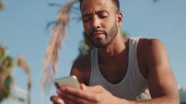 Young bearded male athlete is resting after training, sitting on bench, using smartphone on modern building background