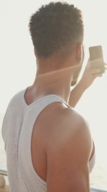 VERTICAL VIDEO: Young bearded male fit athlete taking photo on mobile phone relaxing after workout on the embankment. Back view. Backlight