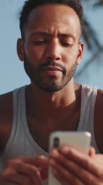 VERTICAL VIDEO: Close up, young bearded male athlete is resting after training, sitting on bench, using smartphone on modern building background