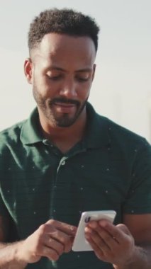 VERTICAL VIDEO: Close up, young smiling man uses phone while sitting in harbor with yachts on background