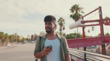 Young smiling man dressed in an olive-colored shirt walks past bridge holding phone in his hands, looks around