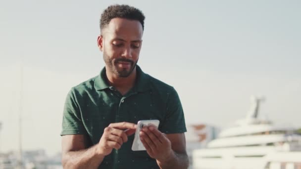 Joven Hombre Sonriente Utiliza Teléfono Mientras Está Sentado Puerto Con — Vídeo de stock