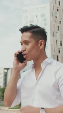 Genç işadamı, modern binaların arka planında akıllı telefondan konuşuyor.