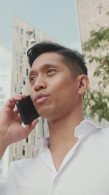 VERTICAL VIDEO: Close-up of young businessman talking on smartphone against modern buildings background