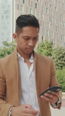 Bej ceketli genç işadamı elinde cep telefonuyla caddede yürüyor.