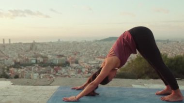 Young woman in bodysuit practices yoga at dawn at viewpoint. Girl raises her hands up