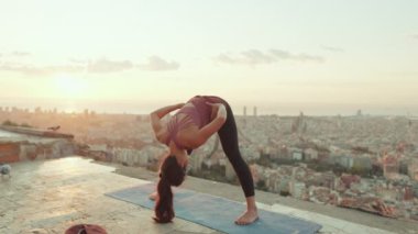 Young woman in bodysuit practices yoga at dawn at viewpoint. Girl raises her hands up