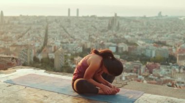 Young woman in bodysuit practices yoga at dawn at viewpoint. Girl raises her hands up