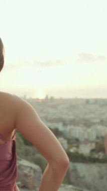 VERTICAL VIDEO: Girl standing at the lookout point at dawn looks at the city. Back view