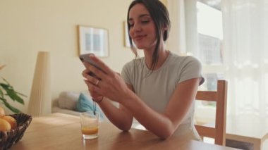 Mutfakta otururken cep telefonuyla fotoğraf çeken gülümseyen genç bir kadın.