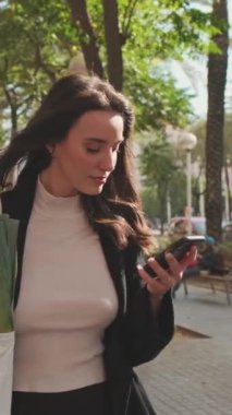 VERTICAL VIDEO, güzel genç bir kadın markette poşetle sokakta yürüyor ve cep telefonu kullanıyor.