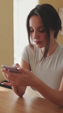 VERTICAL VIDEO, mutfakta otururken cep telefonuyla selfie çeken genç bir kadın.