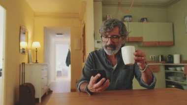 Elderly pleased man drinks coffee from mug and uses mobile phone at home