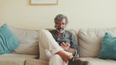 Evde koltukta otururken cep telefonu kullanan yaşlı pozitif bir adam.