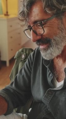VERTICAL VIDEO: Happy senior reading message on his smartphone and drinking coffee while sitting in the kitchen