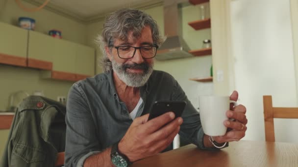 キッチンに座っている間 彼のスマートフォンや飲み物のコーヒーに幸せなシニア読書メッセージ — ストック動画