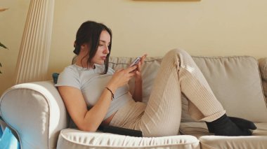 Evdeki kanepede otururken cep telefonu kullanan ve uzaktan kumandayı değiştiren genç bir kadın.