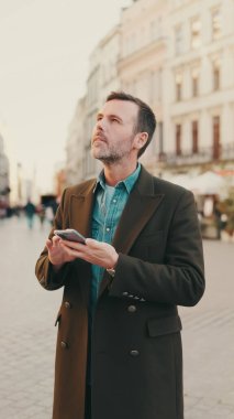 Eski kasabanın arka planında cep telefonunda harita uygulaması kullanan orta yaşlı bir adam.