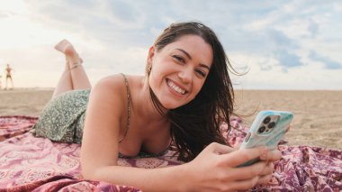 Genç bir kadın sahilde dinleniyor ve cep telefonu kullanıyor.