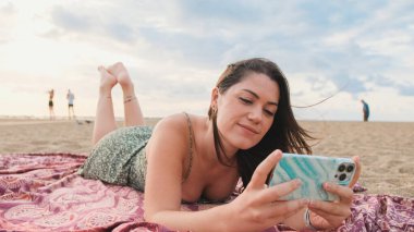 Kumsalda dinlenen ve cep telefonu kullanan gülümseyen genç bir kadın.