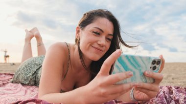 Kumsalda dinlenen ve cep telefonu kullanan gülümseyen genç bir kadın.
