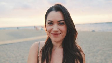 Genç kadın gözlerini açar ve kumsalda dururken kameraya gülümser.