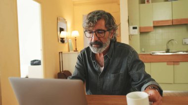 Ev mutfağında oturan yaşlı, pozitif bir adam dizüstü bilgisayarda yazıyor.