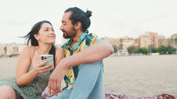 这对年轻夫妇坐在海滩上的时候用手机 背景是建筑物 特写镜头 — 图库照片