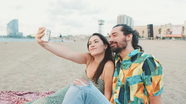快乐的夫妻在沙滩上一边坐着一边用手机自拍 — 图库照片