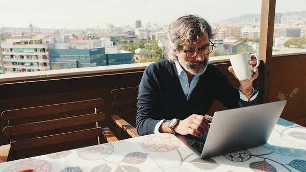 Olgun adam balkonda otururken dizüstü bilgisayarda çalışıyor.