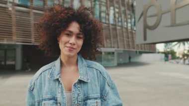 Kız öğrenci üniversite binasının önünde duruyor kameraya bakıyor ve gülümsüyor.