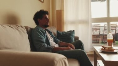 Genç adam oturma odasında oturup televizyon izlerken öğle yemeği yiyor.