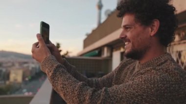 Gülümseyen genç adam balkonda durup fotoğraf çekiyor, cep telefonunda video çekiyor.