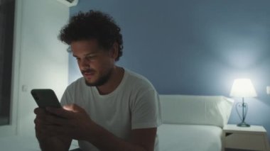 Genç adam yatak odasında oturuyor ve elinde akıllı telefon var.