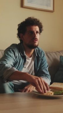 Genç adam oturma odasında oturup televizyon izlerken öğle yemeği yiyor.
