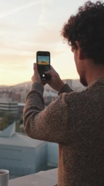Genç adam balkonda duruyor, fotoğraf çekiyor, cep telefonunda video çekiyor.