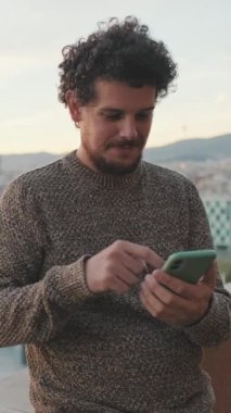 Genç adam balkonda duruyor, cep telefonu kullanıyor.