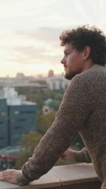 Genç adam balkonda duruyor ve aşağıdaki yola bakıyor.