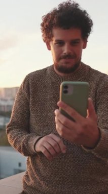 Genç adam balkonda duruyor, cep telefonuyla video görüşmesi yapıyor.