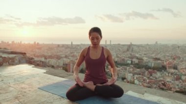 Vücut kıyafetli genç bir kadın güneş doğarken yoga yapıyor. Kız Lotus pozisyonunda otururken ellerini kaldırır.