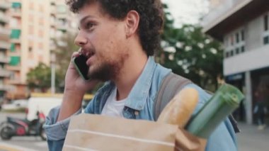 Genç adam yaya geçidinde bir çanta yiyecekle yürüyor, cep telefonuyla konuşuyor.